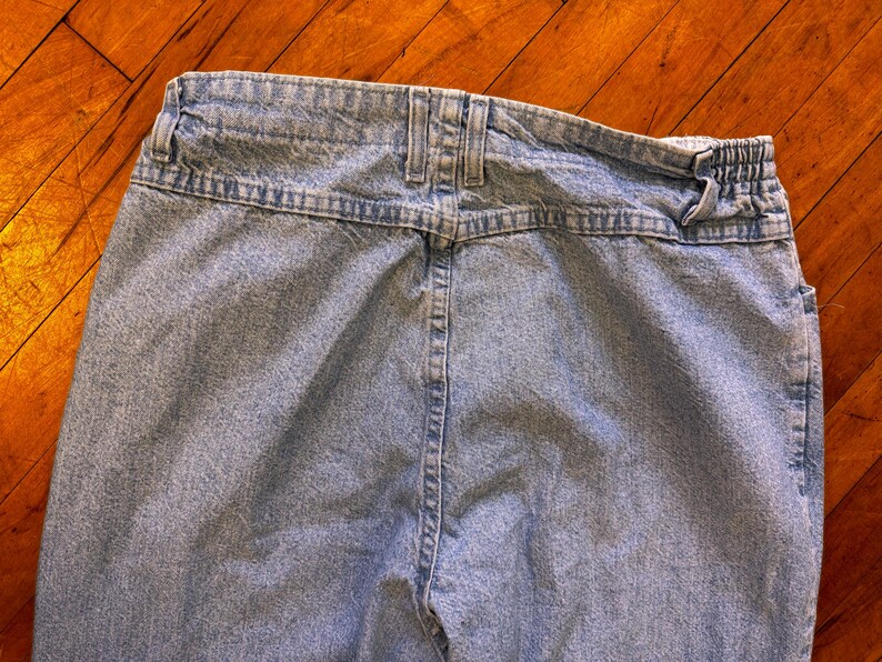 May include: Light blue denim jeans, viewed from the back. The jeans feature a high waist, belt loops, and a partially elastic waistband. The fabric has a faded appearance, with visible stitching details.