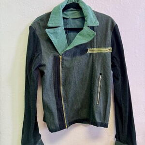 May include: A denim jacket with a teal collar and cuffs, and black corduroy sleeves. The jacket has a diagonal zipper closure and a small zippered pocket on the chest. The jacket is hanging on a black hanger.