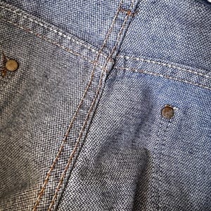 May include: Close-up of denim fabric with visible stitching and metal rivets. The denim has a textured appearance with a subtle herringbone pattern. The stitching is a contrasting color, and the rivets are metallic.