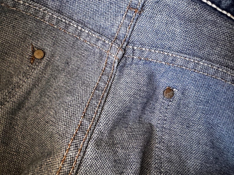 May include: Close-up of denim fabric with visible stitching and metal rivets. The denim has a textured appearance with a subtle herringbone pattern. The stitching is a contrasting color, and the rivets are metallic.