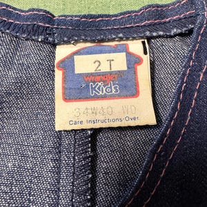 Może przedstawiać: Zbliżenie na dżinsowy element garderoby z metką. Na metce widnieje napis "2T Wrangler Kids" w niebiesko-czerwonym wzorze. R&oacute;żowe przeszycia podkreślają krawędzie ubrania. Tkanina to ciemnoniebieski dżins.