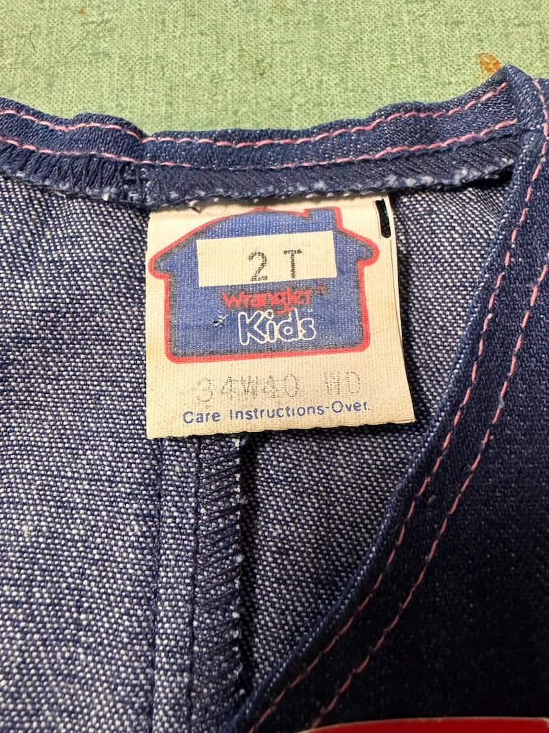 Może przedstawiać: Zbliżenie na dżinsowy element garderoby z metką. Na metce widnieje napis "2T Wrangler Kids" w niebiesko-czerwonym wzorze. R&oacute;żowe przeszycia podkreślają krawędzie ubrania. Tkanina to ciemnoniebieski dżins.