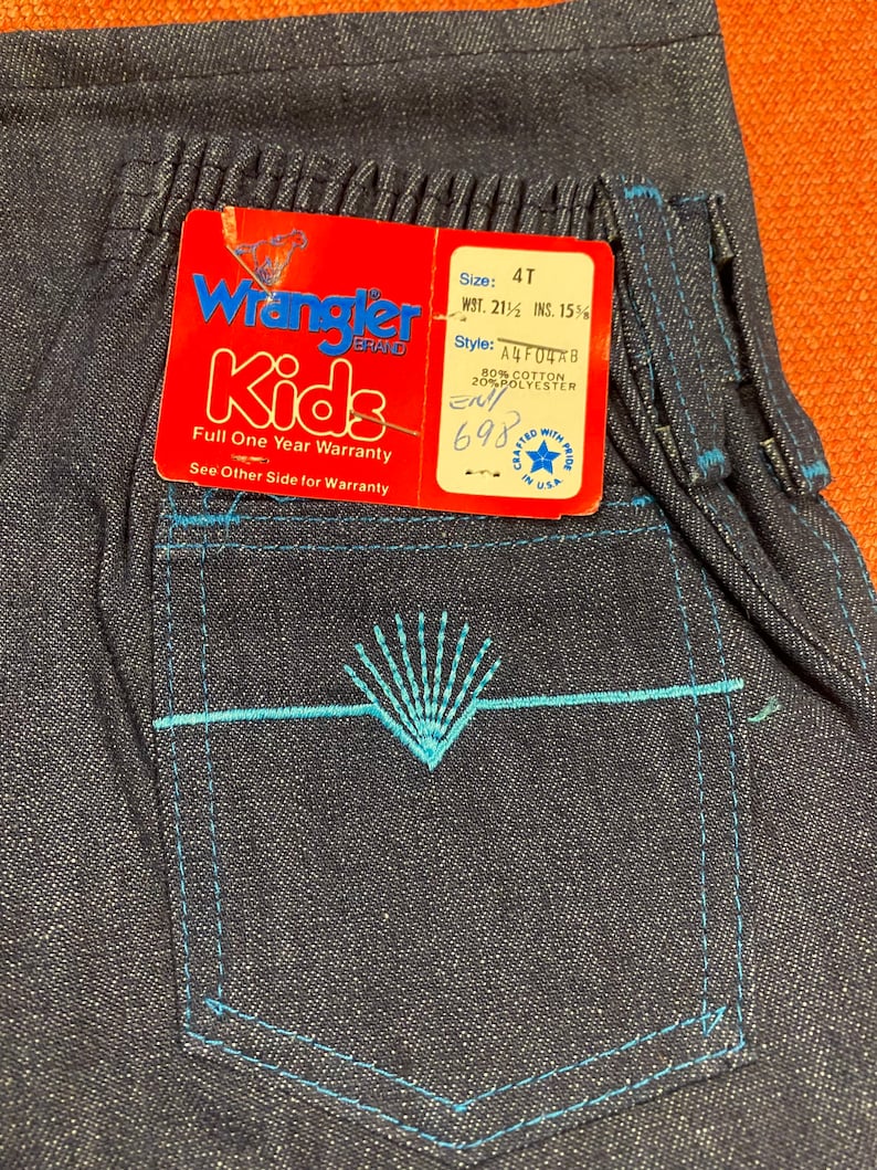 K&ouml;nnte beinhalten: Wrangler Kids blaue Jeans aus Denim mit einem blauen, gestickten Taschendetail. Das Etikett lautet "Wrangler Kids" und enth&auml;lt Gr&ouml;&szlig;en- und Stilinformationen. Das Etikett enth&auml;lt auch die Aufschrift "Volle einj&auml;hrige Garantie" und "Siehe R&uuml;ckseite f&uuml;r die Garantie".