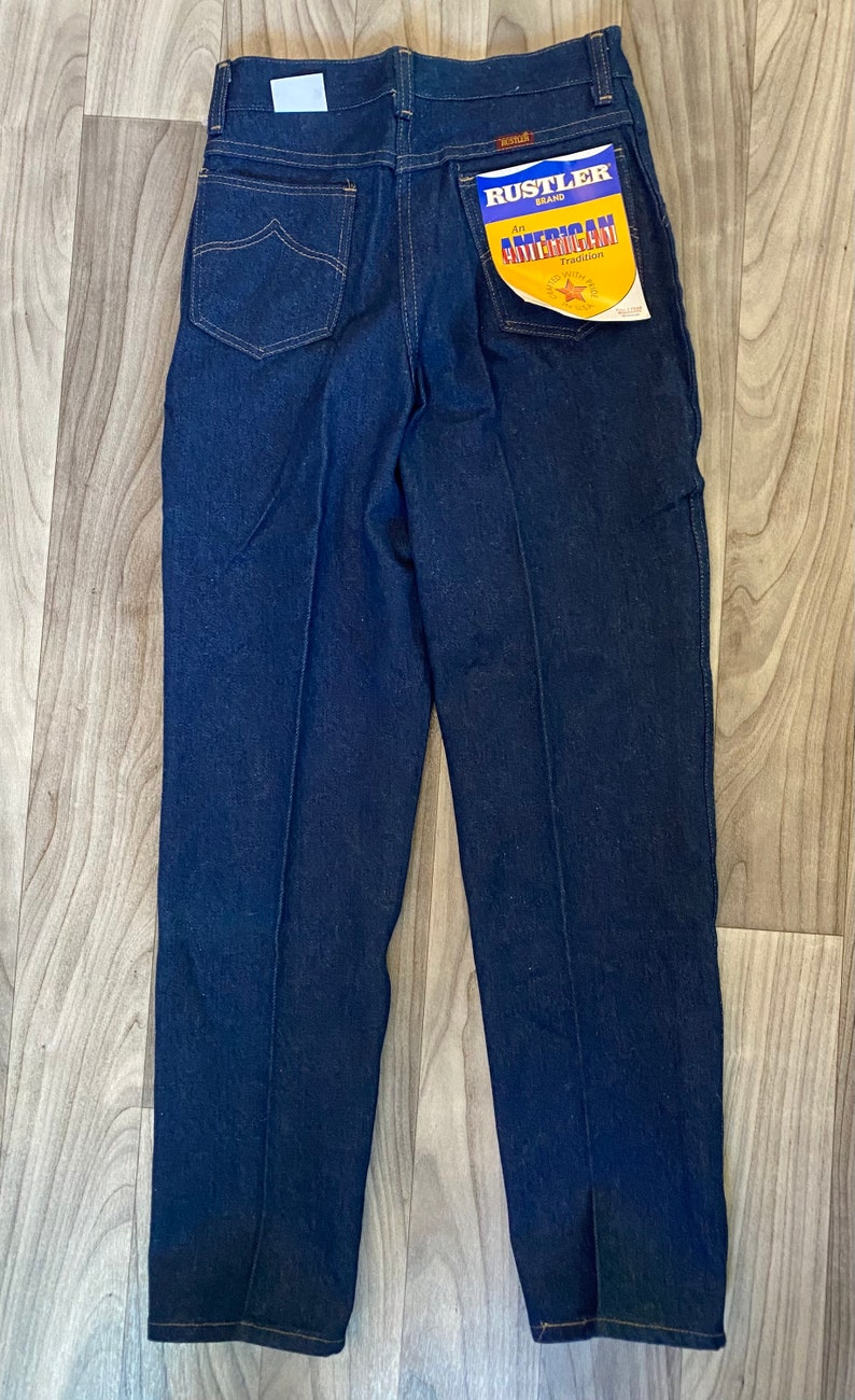 May include: A pair of dark blue denim jeans with a Rustler brand label on the back pocket. The label reads "Rustler Brand An American Tradition."