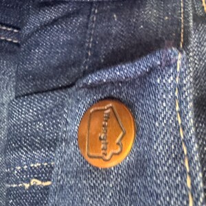 May include: Close-up of a dark blue denim fabric with a copper-colored button. The button has the word "Wrangler" and a house-shaped logo embossed on it. The denim has white stitching.