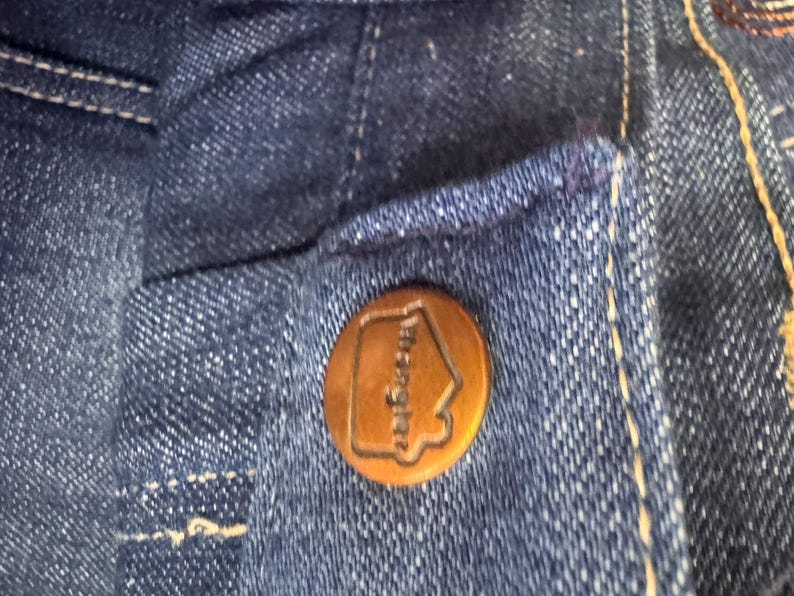 May include: Close-up of a dark blue denim fabric with a copper-colored button. The button has the word "Wrangler" and a house-shaped logo embossed on it. The denim has white stitching.