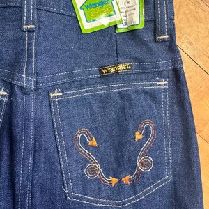 May include: Close-up of vintage Wrangler Girls denim jeans. The jeans are a medium blue wash with white stitching and an embroidered design on the back pocket. The tag reads "Wrangler Girls" and "Size 10".