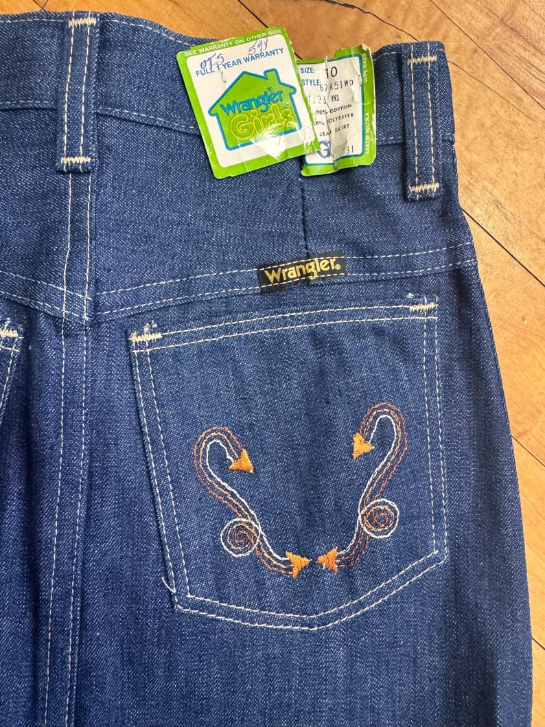 May include: Close-up of vintage Wrangler Girls denim jeans. The jeans are a medium blue wash with white stitching and an embroidered design on the back pocket. The tag reads "Wrangler Girls" and "Size 10".