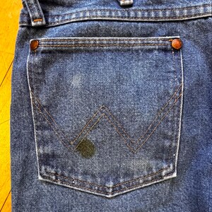 May include: Close-up of the back pocket of blue denim jeans. The pocket has a classic design with a stitched "W" shape and is secured with copper rivets. The denim shows signs of wear and a dark stain.