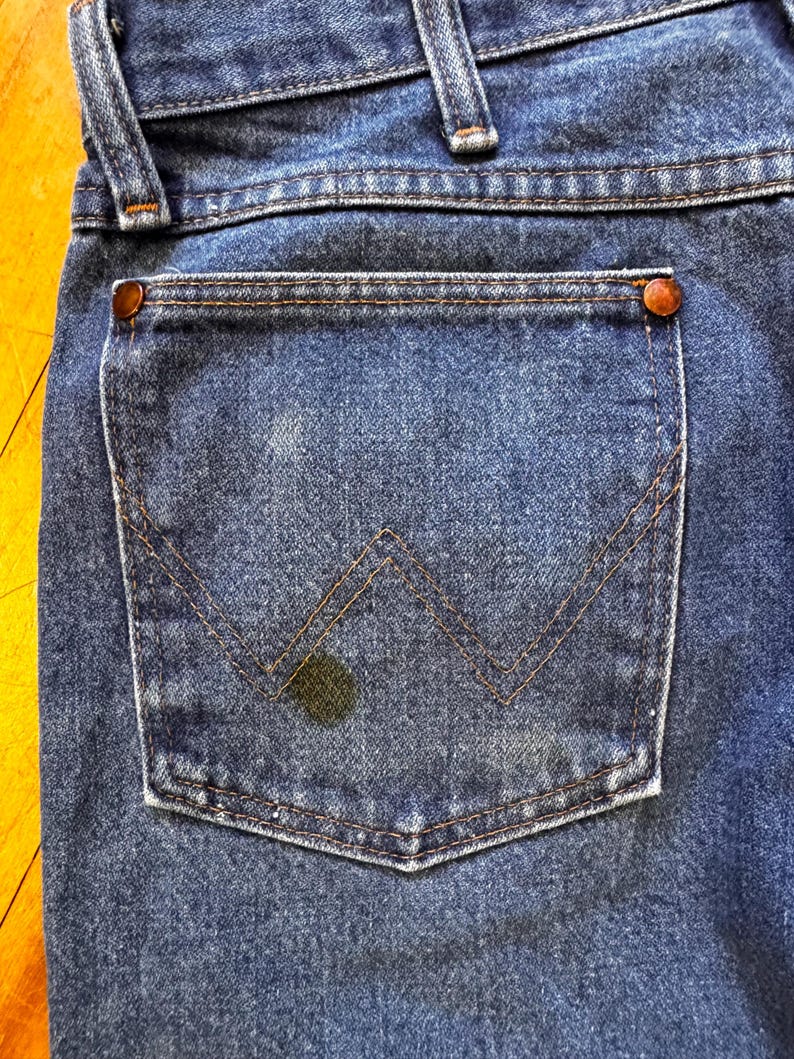May include: Close-up of the back pocket of blue denim jeans. The pocket has a classic design with a stitched "W" shape and is secured with copper rivets. The denim shows signs of wear and a dark stain.
