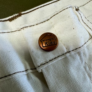 K&ouml;nnte beinhalten: Nahaufnahme einer wei&szlig;en Hose mit einem kupferfarbenen Knopf, auf dem "Wrangler Kids" steht.