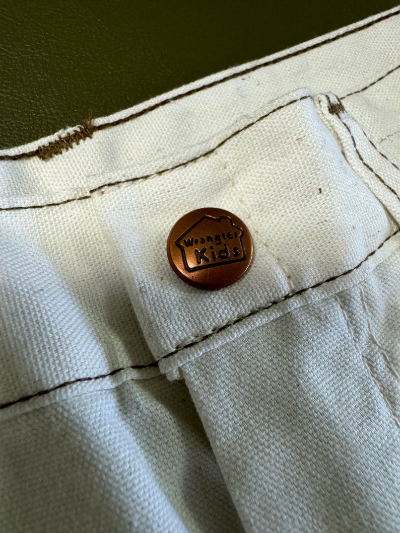 K&ouml;nnte beinhalten: Nahaufnahme einer wei&szlig;en Hose mit einem kupferfarbenen Knopf, auf dem "Wrangler Kids" steht.