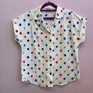 May include: A white short-sleeve button-down shirt featuring a repeating pattern of multicolored polka dots in red, blue, green, black, and tan, interspersed with small black "x" marks. The shirt has a classic collar and cuffed sleeves.