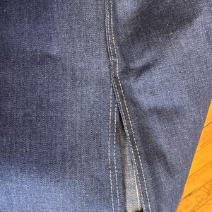 May include: Close-up of dark blue denim fabric with visible texture and white stitching. The image shows a split seam detail, highlighting the construction of the garment. The denim has a classic, worn look.