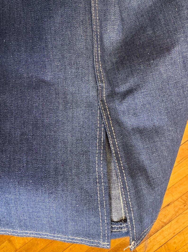 May include: Close-up of dark blue denim fabric with visible texture and white stitching. The image shows a split seam detail, highlighting the construction of the garment. The denim has a classic, worn look.