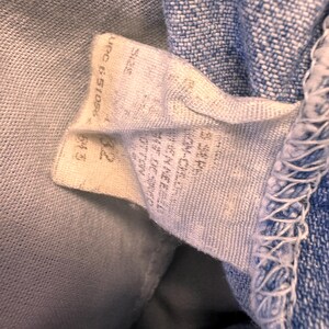 May include: Close-up of a light blue denim fabric with a white tag attached. The tag has printed text, including a size indication of "32" and a UPC code. The denim has visible stitching along the edge.