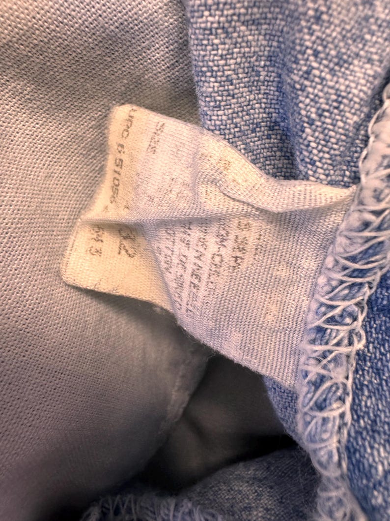 May include: Close-up of a light blue denim fabric with a white tag attached. The tag has printed text, including a size indication of "32" and a UPC code. The denim has visible stitching along the edge.