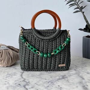 May include: A dark green crocheted handbag with a wooden top handle and a green and gold chain strap. The bag has a small tag that says "handmade".