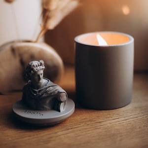Könnte beinhalten: Eine graue Kerze mit einer weißen Flamme steht auf einer Holzoberfläche neben einer kleinen grauen Büste von Apollo. Die Büste steht auf einem runden Sockel mit der Aufschrift "Apollo".