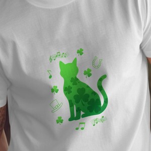 Peut inclure: T-shirt blanc avec une silhouette de chat vert remplie de trèfles. Le motif comprend des notes de musique, un fer à cheval et un chapeau haut de forme, tous en vert. Un t-shirt graphique sur le thème de la Saint-Patrick.