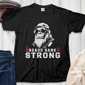 Peut inclure: T-shirt noir avec un gorille portant des lunettes de soleil et une casquette, avec le texte "BEARD GAME STRONG" en blanc. Des flammes encadrent le texte. L'image comprend un jean, une veste et des bottes.