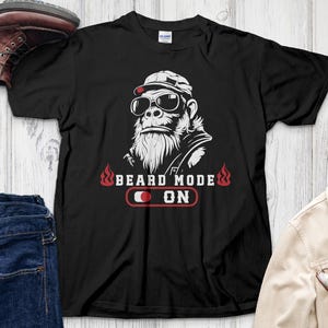 May include: Black t-shirt with a graphic of a gorilla wearing sunglasses and a cap, with a long white beard. The text "BEARD MODE ON" is above a red and white design. Brown leather boots and blue jeans are also visible.