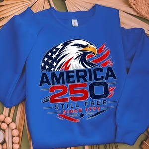 Peut inclure: Un sweat-shirt bleu roi avec un aigle chauve américain et le texte "AMERICA 250 STILL FREE SINCE 1776". Le motif de l'aigle reprend les couleurs du drapeau américain.