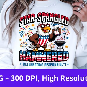 May include: White sweatshirt with a patriotic design featuring an eagle wearing sunglasses and holding a can of "Freedom Brew". The text reads "Star-Spangled Hammered" with fireworks and a grill. The phrase "Celebrating Responsibly!" is also included.