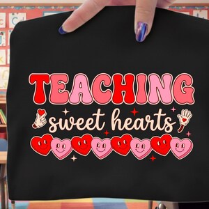 Peut inclure: T-shirt noir avec l'inscription "TEACHING sweet hearts" dans un style rétro. Le motif comprend un geste de la main en forme de cœur, une main avec un cœur et des cœurs souriants. Le t-shirt est tenu par une main.