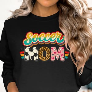 Peut inclure: Sweat-shirt noir avec l'inscription "Soccer MOM" dans un style rétro. Le mot "Soccer" est en arc-en-ciel, et "MOM" présente des ballons de foot, un imprimé léopard et des nœuds roses. Un motif à rayures colorées est de chaque côté.