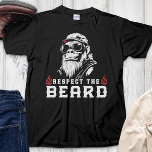 Puede incluir: Camiseta negra con un gráfico de un gorila con gafas de sol y un sombrero, con el texto "Respect the Beard" en blanco. La camiseta se muestra con botas marrones, vaqueros azules y una camisa a cuadros.