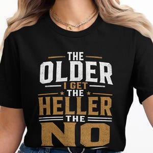 Peut inclure: T-shirt noir avec l'inscription "THE OLDER I GET THE HELLER THE NO" en blanc et or. Le design est centré sur le devant. Le t-shirt a un col rond et des manches courtes, idéal pour un look décontracté.