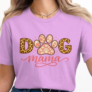 Peut inclure: T-shirt lilas avec l'inscription "DOG Mama". Le mot "DOG" est imprimé en léopard, avec une empreinte de patte au centre. L'empreinte de patte est rose clair avec des formes de cœur. Vêtement décontracté.