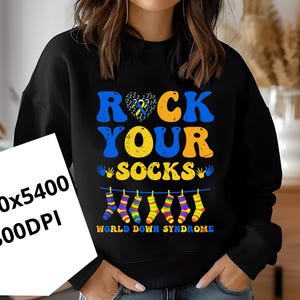 Peut inclure: Sweat-shirt noir avec l'inscription "ROCK YOUR SOCKS" en bleu et jaune. Un motif en forme de cœur avec des pièces de puzzle bleues et jaunes est au-dessus du texte. En dessous, des chaussettes colorées sont suspendues à une corde à linge. Le texte "WORLD DOWN SYNDROME" est en dessous.