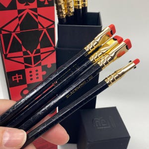 May include: Black pencils with gold-coloured metal ferrules and red erasers. The pencils are black with a diamond pattern and the text "BLACKWING 20". A black box with a red and black design is visible in the background.