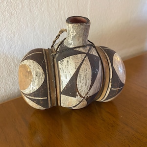 Puede incluir: Un recipiente de cerámica con forma redondeada de varios lóbulos y un cuello central. La superficie presenta un acabado mate y desgastado con patrones geométricos en negro, blanco y tostado. Un cordel fino y oscuro rodea el cuello y el cuerpo.
