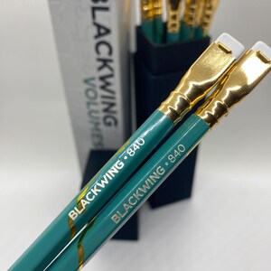 以下が含まれることがあります： 金色のフェルールと白い消しゴムが付いたティール色の鉛筆2本。鉛筆には「BLACKWING 840」という白い文字が刻印されています。背景には、追加の鉛筆が入った黒い箱と「BLACKWING VOLUMES」と書かれた白い箱が見えます。