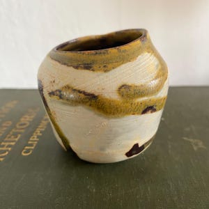 Może przedstawiać: Mały, ręcznie robiony ceramiczny wazon o unikalnym, organicznym kształcie. Wazon ma kremową podstawę z wirującymi wzorami w kolorze musztardowej żółci i ciemnobrązu. Wnętrze jest w kolorze ciemnobrązowym.