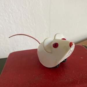 May include: A white toy mouse with red eyes and a pink tail. The mouse has black wheels and is sitting on a red surface. The toy is a vintage item.