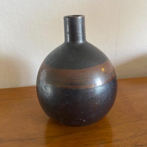 May include: A spherical ceramic vase with a narrow neck, featuring a dark charcoal black finish and a prominent horizontal band of earthy brown glaze encircling the centre of the rounded body, creating a smooth and reflective surface texture.