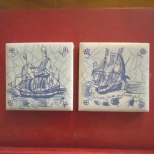 May include: Two square ceramic tiles with a blue and white nautical design. Each tile features a detailed illustration of a sailing ship on the water. The tiles have a decorative border and are set against a red background.