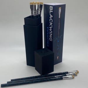 May include: A collection of Blackwing Volumes pencils and accessories. The image shows a set of pencils, a pencil sharpener, and a box with the text "Blackwing Volumes". The pencils have a black and blue marbled design.