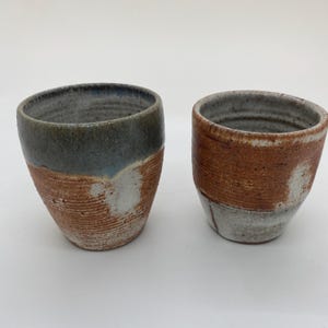 Könnte beinhalten: Zwei handgefertigte Keramikbecher. Die Becher haben ein strukturiertes, erdiges Aussehen mit einem Farbschema aus Braun, Grau und Weiß. Der obere Teil eines Bechers ist graublau, während der andere braun ist. Die unteren Abschnitte sind eine Mischung aus Braun und Weiß.