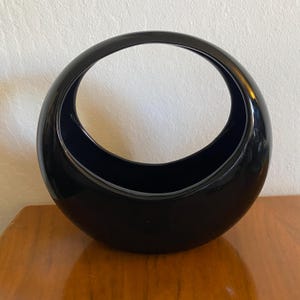 May include: A black ceramic vase with a unique crescent moon shape and a large opening at the top. The vase is glossy and has a smooth finish.