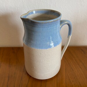 Puede incluir: Una jarra de cerámica blanca con un esmalte azul claro en la mitad superior. La jarra tiene un cuerpo redondeado y un asa.