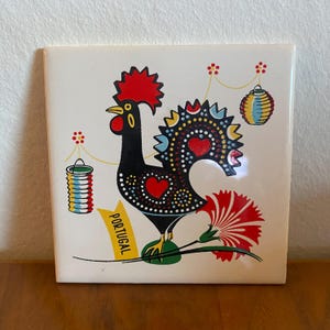 May include: Square ceramic tile featuring a stylized black rooster with a red comb and heart-shaped details. The rooster is adorned with colorful accents and is holding a red flower. The tile has the word "PORTUGAL" on a yellow banner.