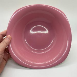 Op de afbeelding: Een ronde, roze keramische kom met een glanzende afwerking. De kom heeft een breed, ondiep ontwerp en een glad interieur. De kom wordt door een hand vastgehouden en de achtergrond is wit.