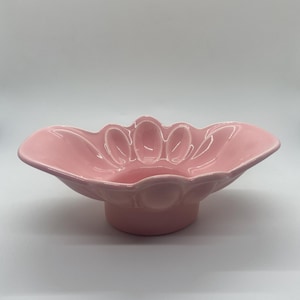 May include: A pink ceramic bowl with a scalloped edge and a raised centre design. The bowl has a unique shape and is perfect for serving snacks or holding small items.