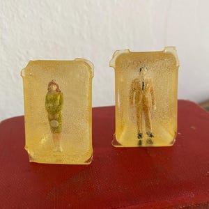 May include: Two small, rectangular resin sculptures. One features a woman in a green dress, the other a man in a suit. Both figures are encased in a translucent, amber-colored resin. The sculptures are set on a red surface.