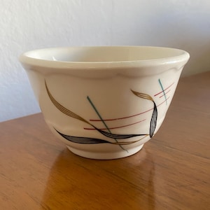 May include: Cream-colored ceramic bowl with a scalloped rim. The bowl is decorated with a mid-century modern design featuring brown, black, red, and teal abstract lines and leaf-like shapes. The bowl sits on a wooden surface.